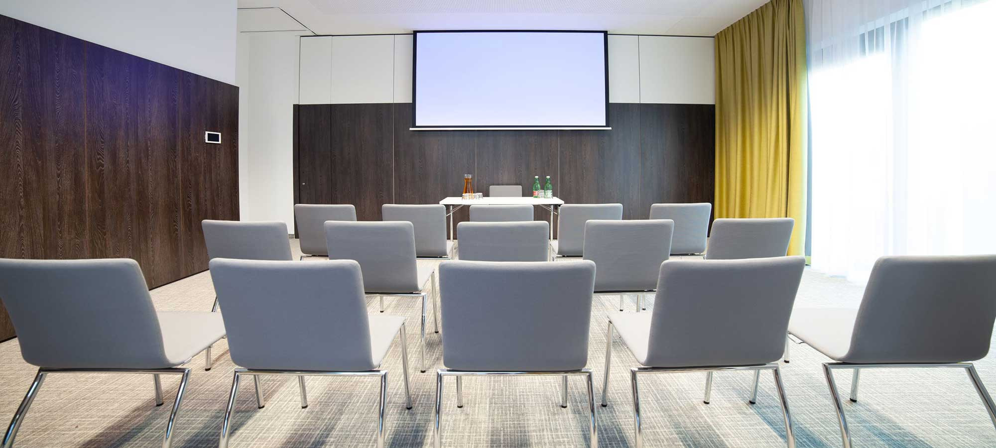 Kleiner Konferenzraum mit grauen Stühlen, weißem Tisch, Flaschen, Gläsern, Leinwand, Holzverkleidung und gelbem Vorhang.