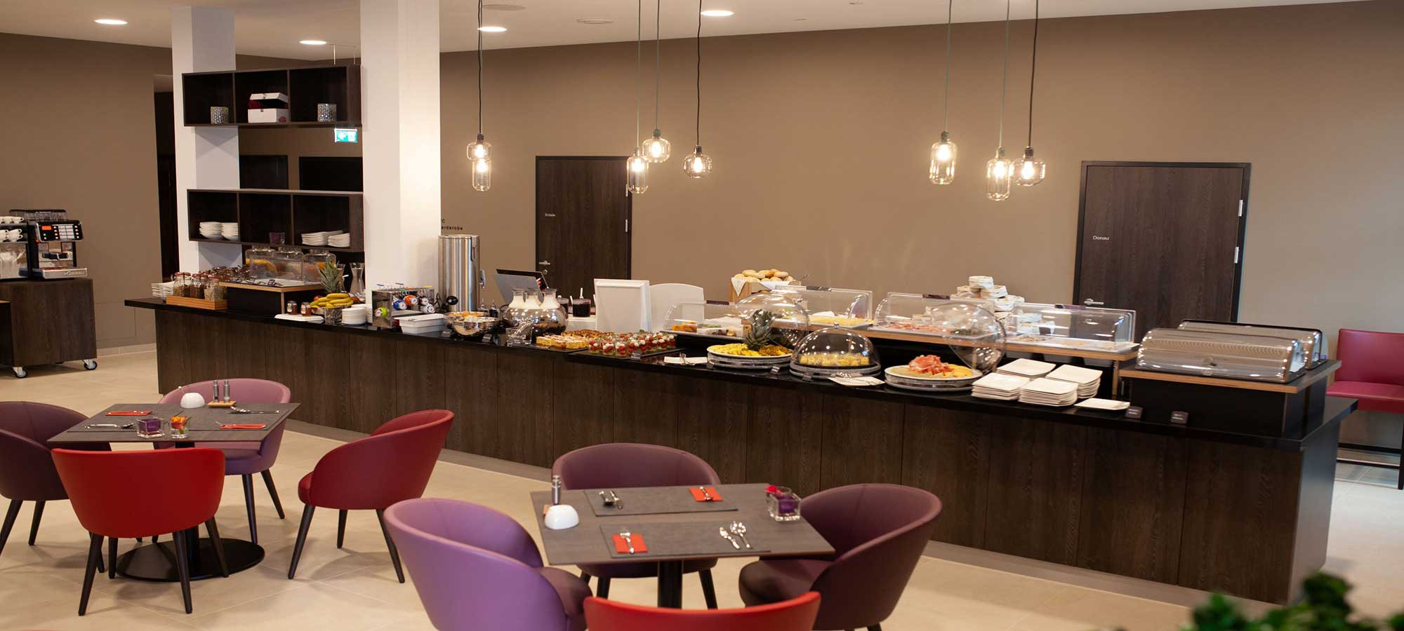 Modernes Café mit langer Theke, Speisen, Getränken und bunten Sitzbereichen in warm beleuchtetem Ambiente.