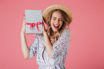 Person mit Strohhut und Blumenoutfit hält Geschenk mit roter Schleife vor rosa Hintergrund